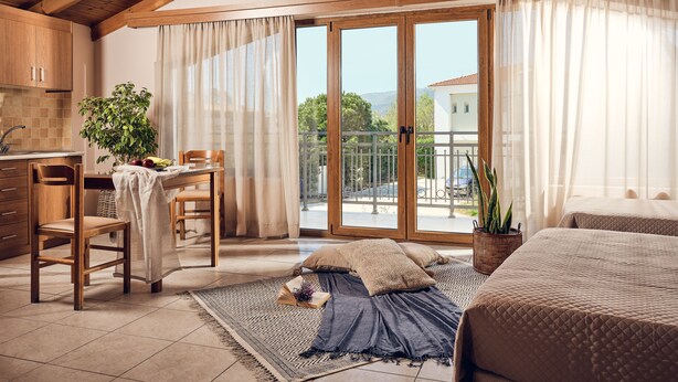 A cozy room with a wooden ceiling, a dining area with a table and chairs, a bed, and a balcony with a view of the countryside.