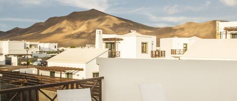 A terrace with two white lounge chairs and a small table, overlooking a mountainous landscape and white buildings.