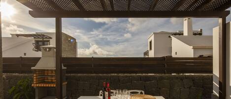 An outdoor patio with a table set for dining, white chairs, a wicker basket, wine bottles, and glasses.