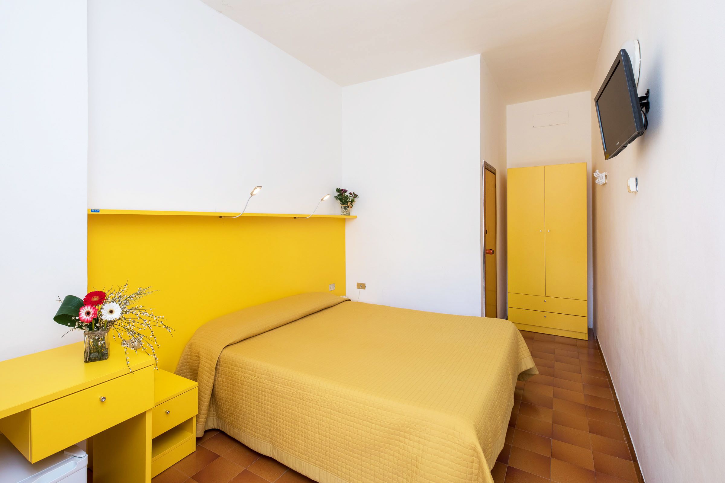 Una habitación con una cama, un escritorio amarillo, un televisor montado en la pared y una puerta que da a otra habitación.