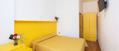 Una habitación con una cama, un escritorio amarillo, un televisor montado en la pared y una puerta que da a otra habitación.
