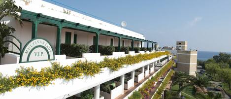 A hotel with a long white facade, green accents, and a balcony with yellow flowers.