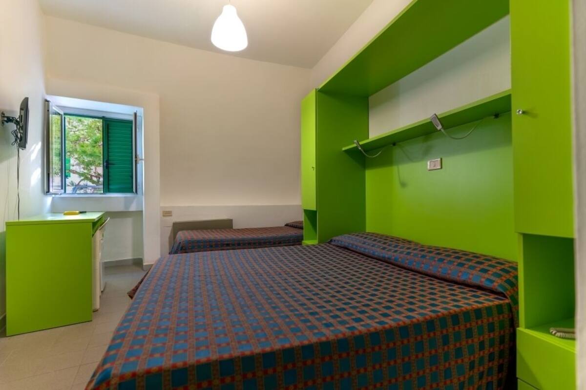 A room with two beds, a desk, and a window with green shutters.
