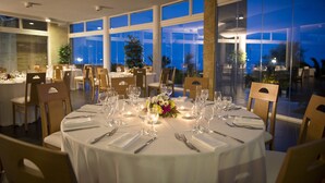 A dining area with round tables set for a meal, large windows, and indoor plants. - Cala Corvino Hotel Resort (Monopoli)