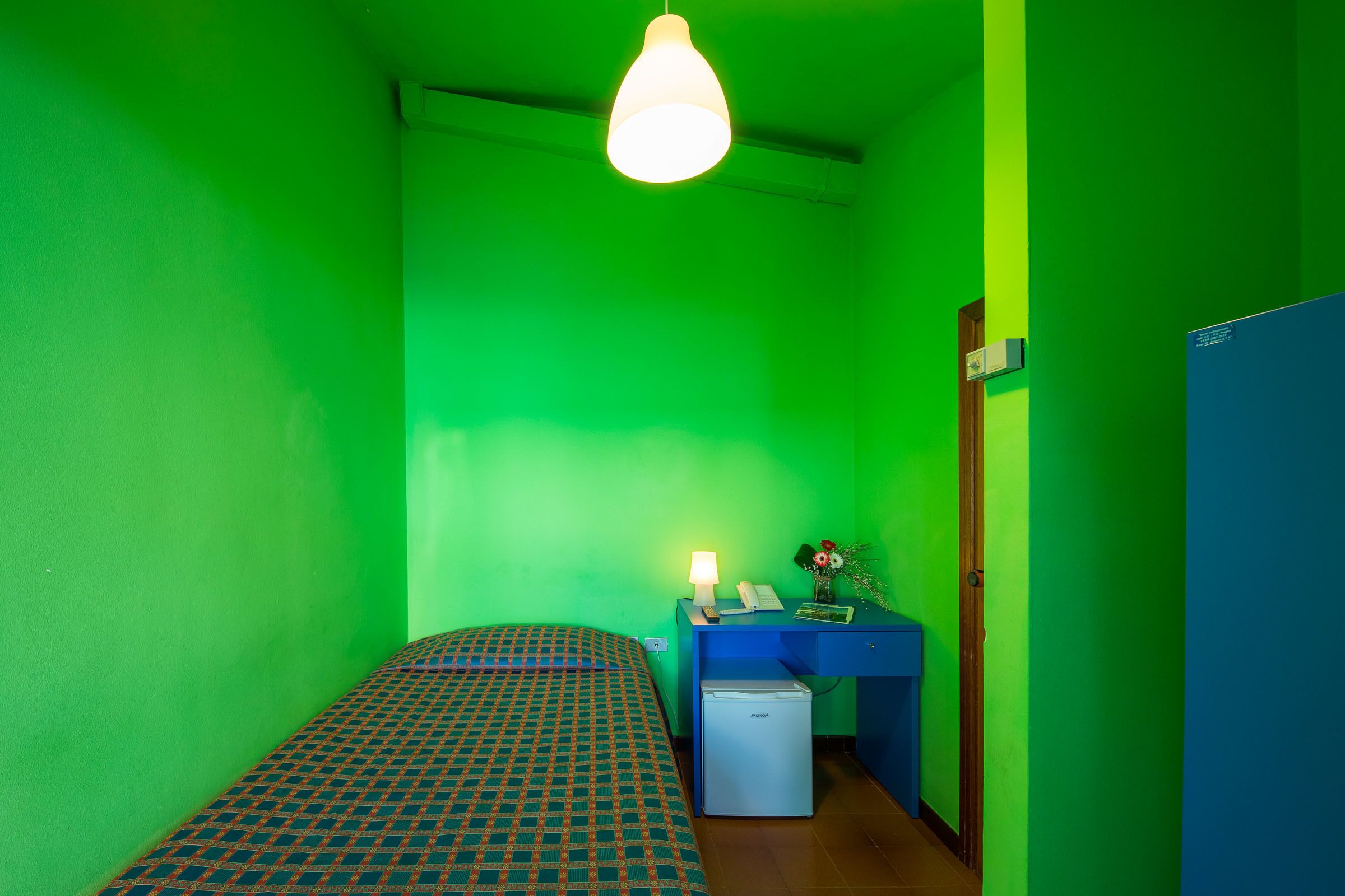A small room with a bed, a desk, a fridge, and a door.