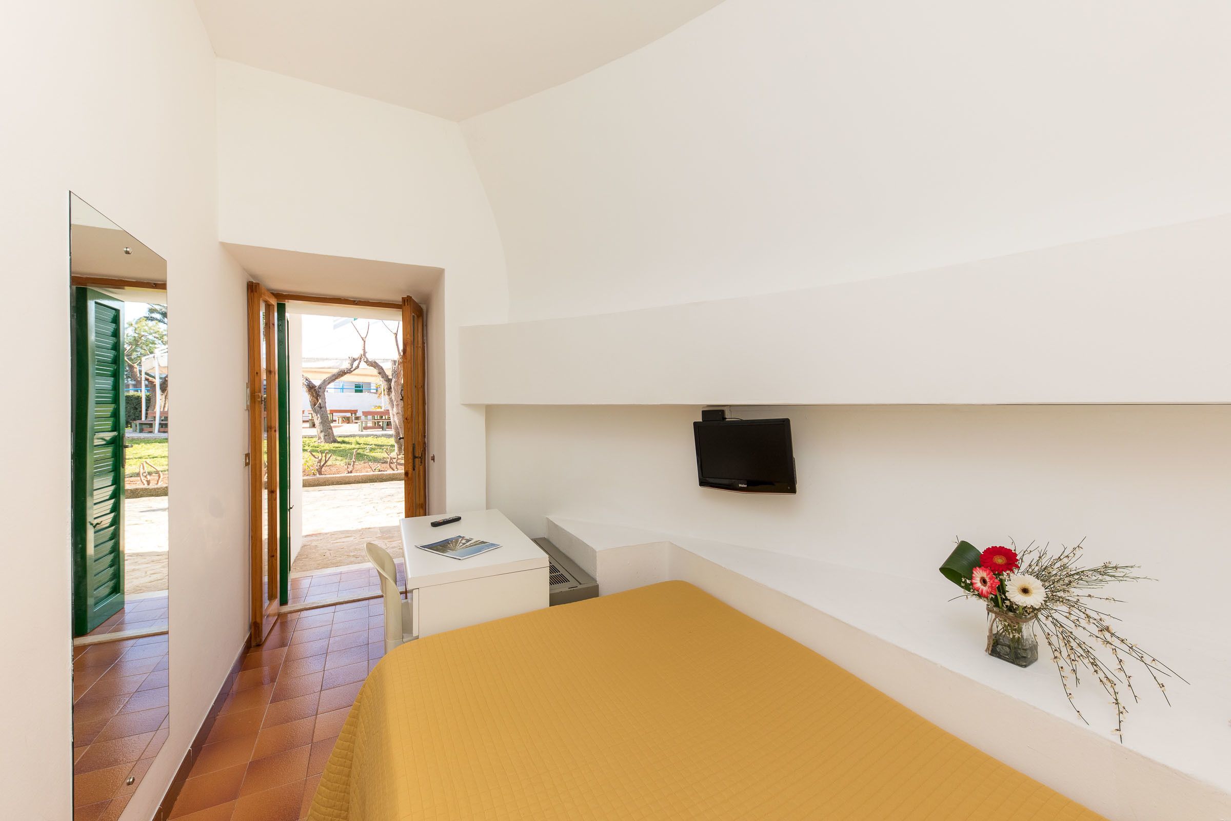Un dormitorio con cama, escritorio, silla, televisor montado en la pared y ventana con vistas al exterior.