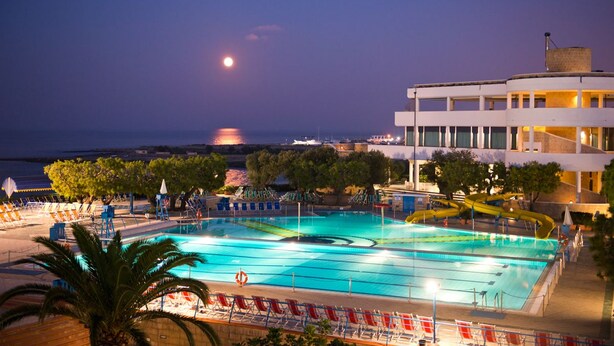 A swimming pool with multiple lanes, surrounded by lounge chairs and umbrellas, and a building with a balcony overlooking the pool. - Cala Corvino Hotel Resort (Monopoli)