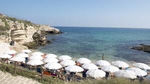 A coastal beach with white umbrellas, rocky cliffs, and clear blue water. - Cala Corvino Hotel Resort (Monopoli)