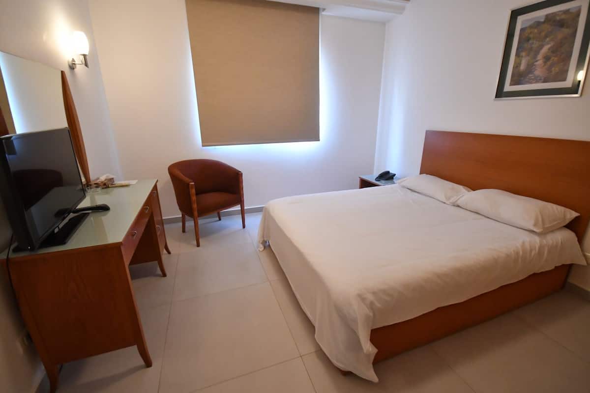 Hotellihuone, jossa on sänky, työpöytä tietokoneella, tuoli, yöpöytä ja ikkuna.
