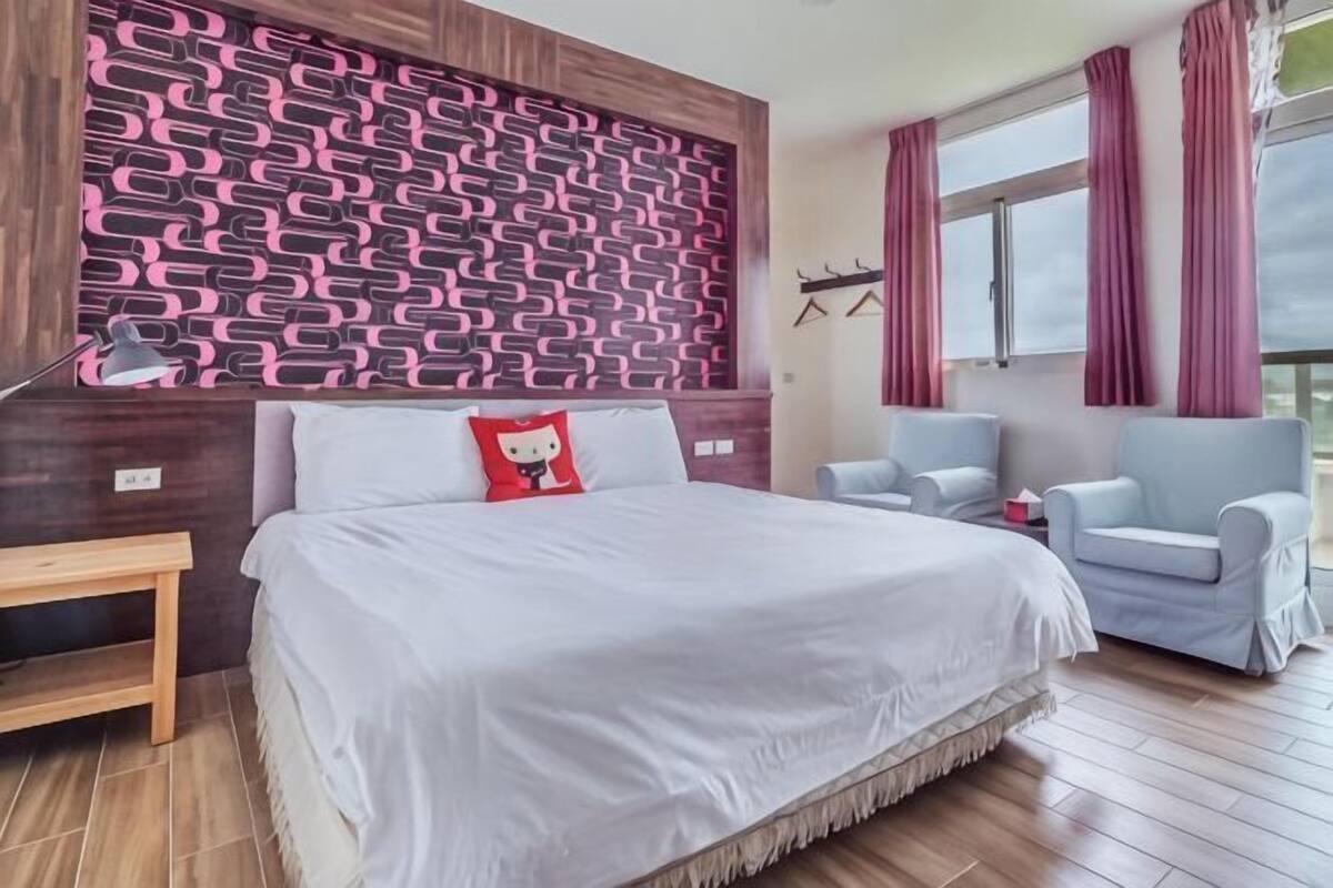 A bedroom with a patterned wall, a bed, two chairs, a nightstand, and a window with curtains.
