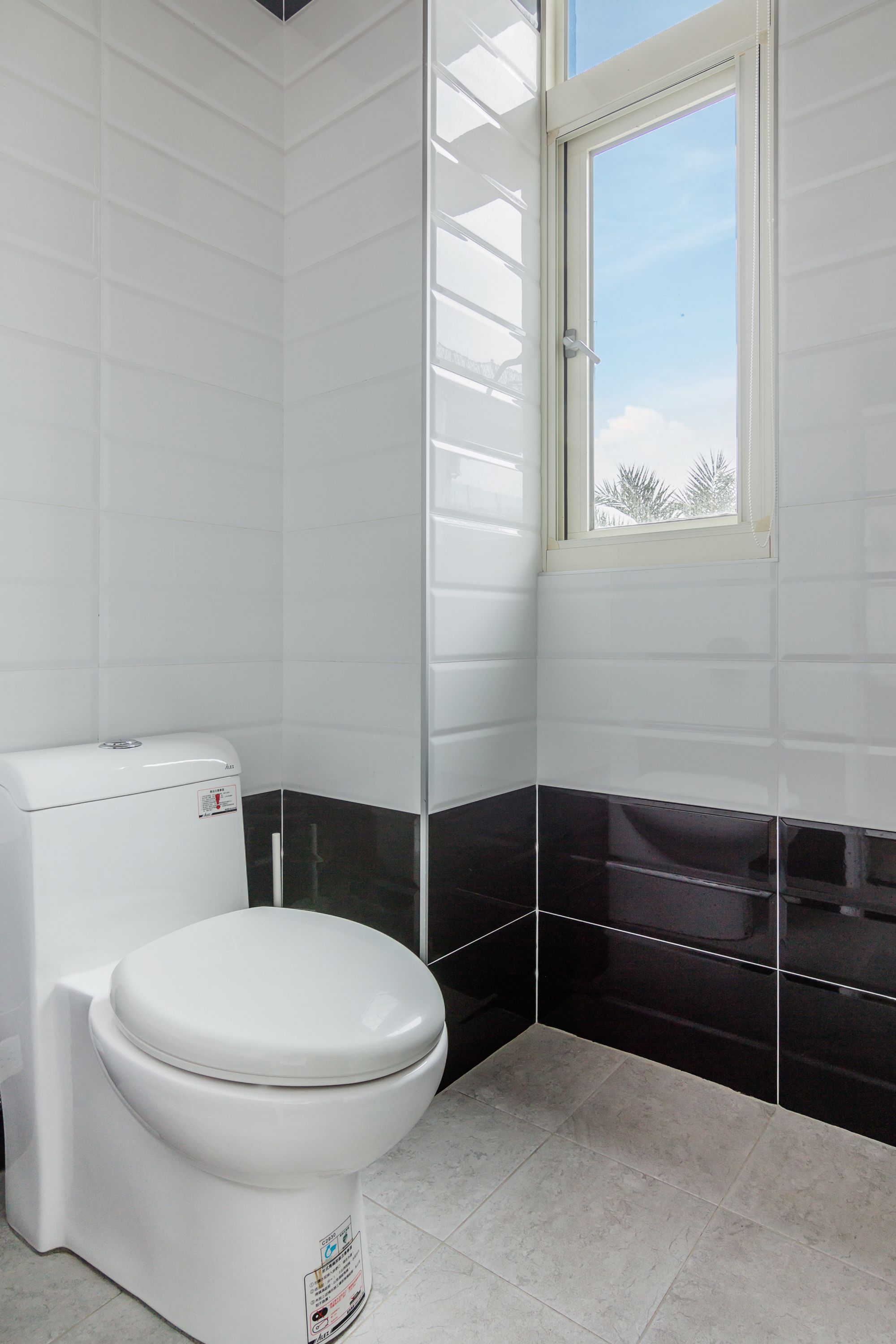 Un baño con paredes cubiertas de azulejos blancos, una sección inferior negra, un inodoro blanco y una ventana con vistas a palmeras.