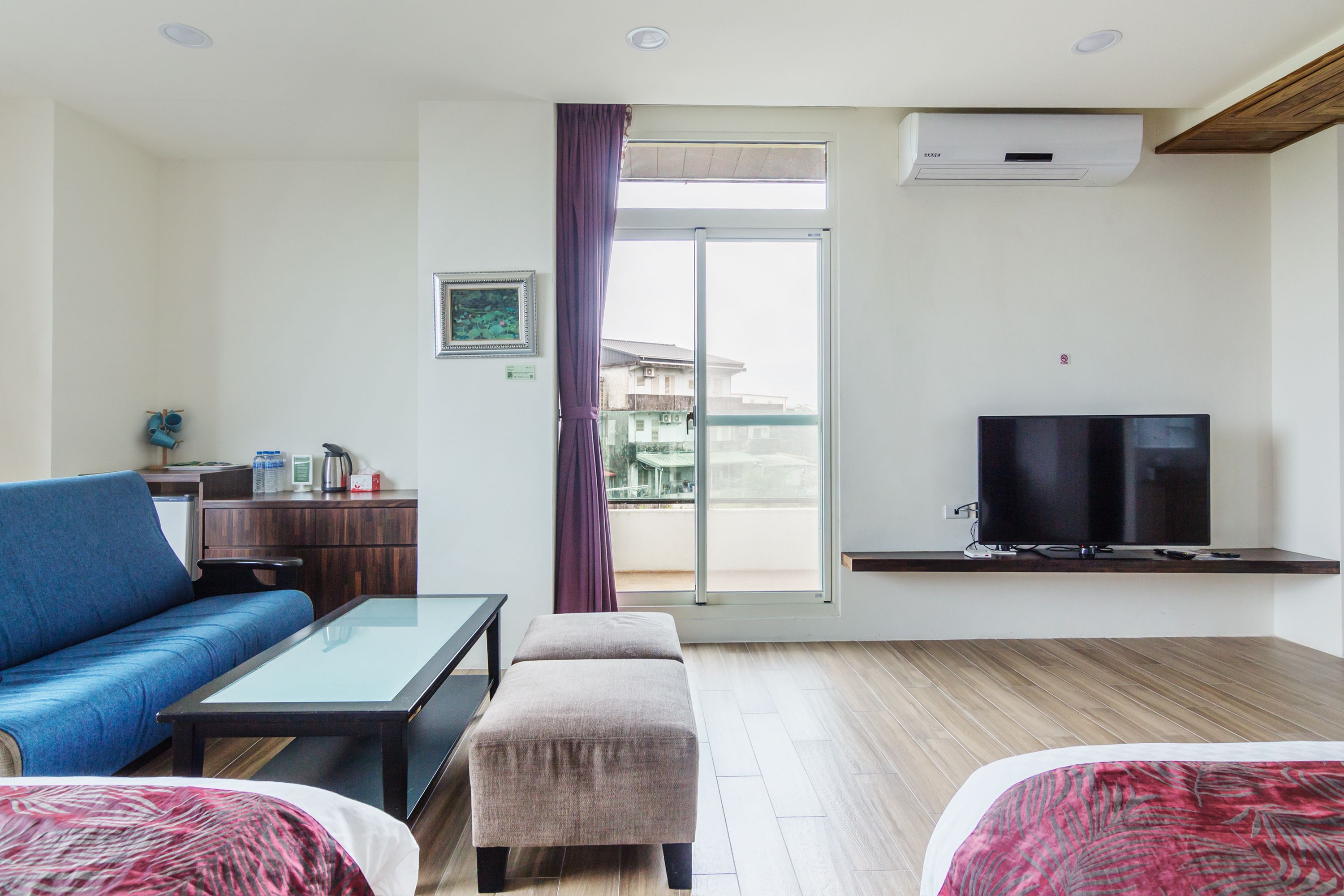 Habitación de hotel con sofá, una mesita, un reposapiés, televisor sobre un mueble, puerta corrediza de vidrio al balcón y un área de kitchenette.
