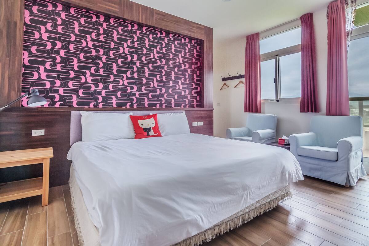 A modern hotel room with a large bed, two armchairs, a wooden nightstand, and a patterned headboard.