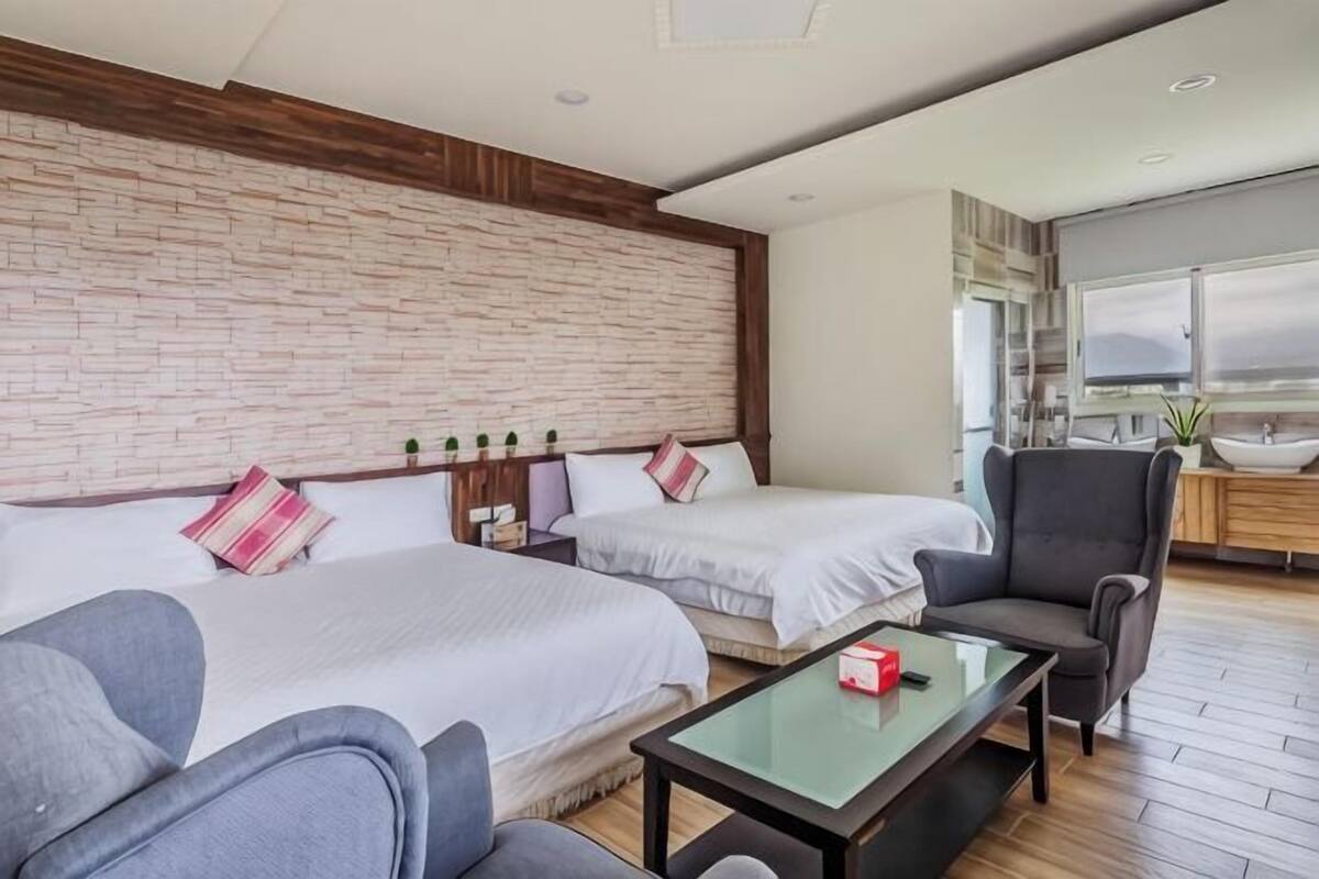 A hotel room with two beds, a sofa, a small table, a chair, a brick wall, a window, and a bathroom with a sink.