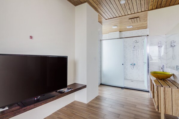Un baño moderno con un espejo grande, un tocador de madera, una cabina de ducha de vidrio y un televisor de pantalla plana montado en la pared.