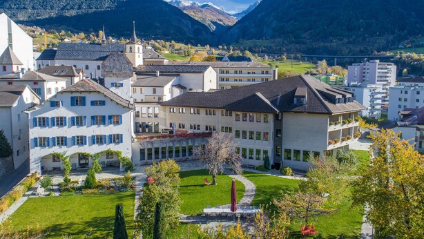 A large building with multiple floors, a courtyard with a fountain, and a mountainous backdrop. - Gästehaus St. Ursula (Brig)