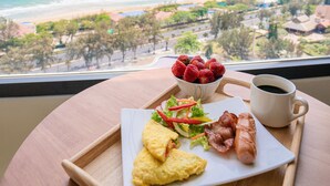 A breakfast tray with scrambled eggs, bacon, strawberries, and a cup of coffee, viewed from a window overlooking a beach.