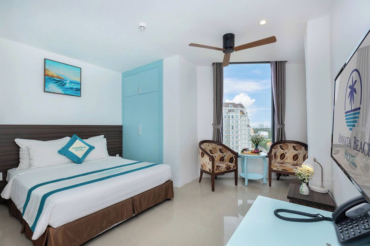 A hotel room with a bed, a desk with a phone, two chairs, a ceiling fan, a window with a view of buildings, and a painting on the wall.