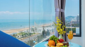 A room with a large window overlooking a beach and cityscape, a table with fruits and a drink, and a vase with flowers.