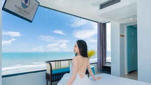 A room with a large window overlooking the beach, a bed, a chair, and a sign for Annata Beach Hotel.