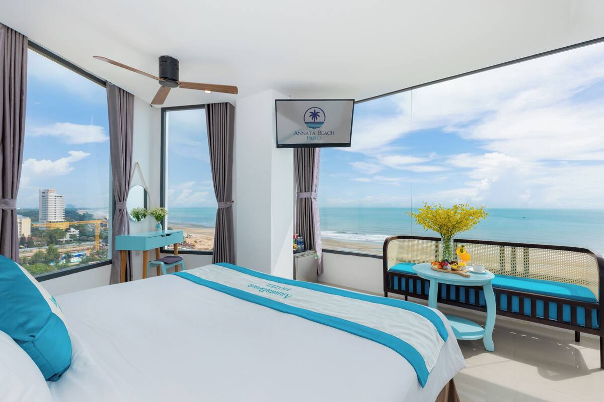 A hotel room with a bed, a desk, a chair, a small table with a vase of flowers, and a balcony with a view of the beach and ocean.