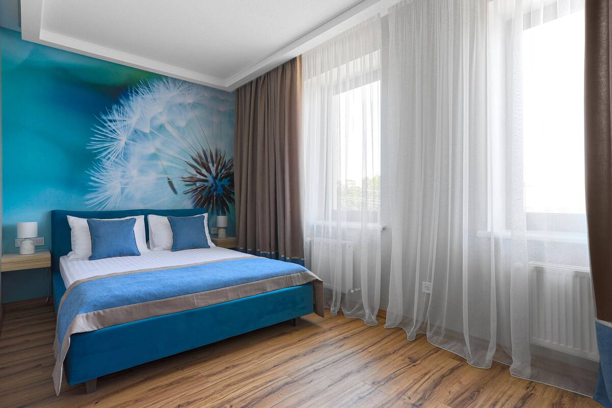 A bedroom with a bed, a nightstand, a lamp, a window with sheer curtains, and a wall mural.