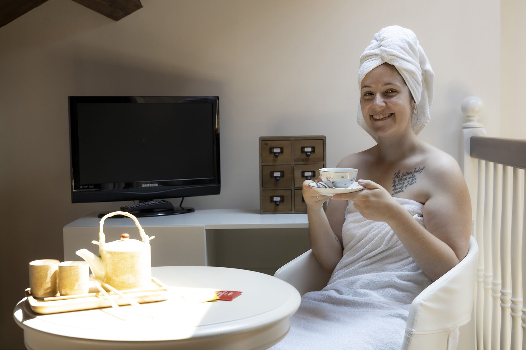 Eine Person in einem weißen Bademantel sitzt an einem Tisch mit Tasse und Untertasse, einem Teekessel und einem Tablett mit Tassen und Teekanne. Ein Fernseher ist an der Wand dahinter angebracht, und zur Rechten befinden sich Aufbewahrungsboxen an der Wand.