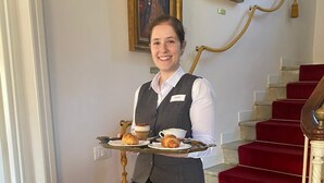 Un membre du personnel de l’hôtel, portant un plateau avec des croissants et du café, se tient devant un escalier recouvert d’un tapis rouge et orné de rampes dorées.