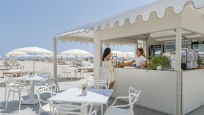 An outdoor cafe with white tables and chairs, a counter with a beverage dispenser, and umbrellas in the background. - Residenza Parco Fellini (Rimini)