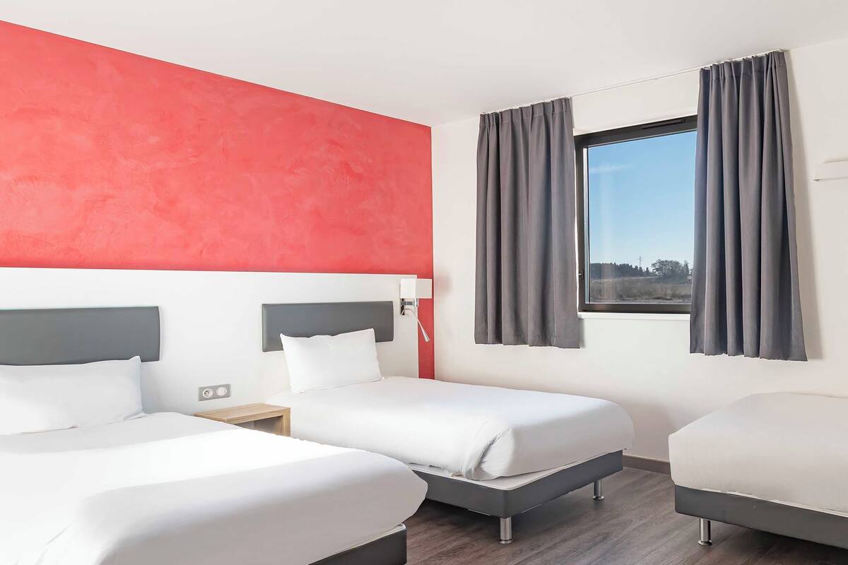 Ein Hotelzimmer mit drei Betten, einer roten Akzentwand, einem Fenster mit Vorhängen und Blick ins Freie.
