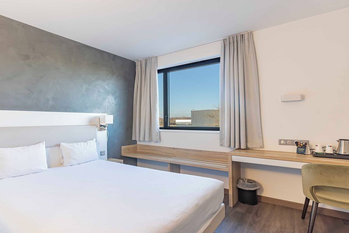 Ein Hotelzimmer mit Bett, Schreibtisch, Stuhl, einem Fenster mit Vorhängen und Blick nach außen.