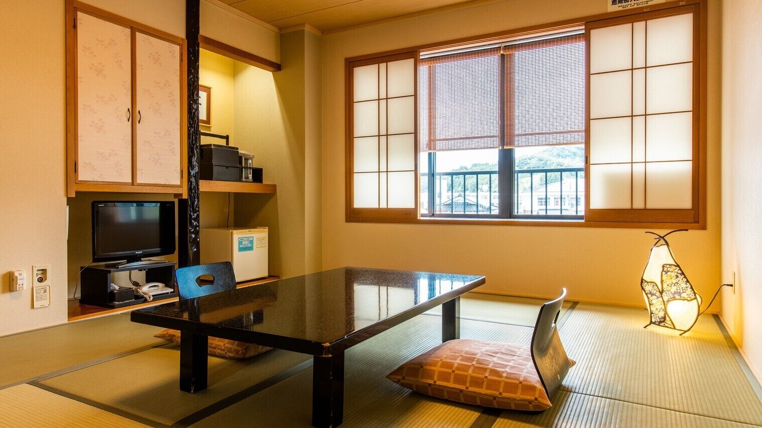 Quarto japonês tradicional com tatames, uma mesa baixa, televisão, frigorífico e janela com painéis shoji.