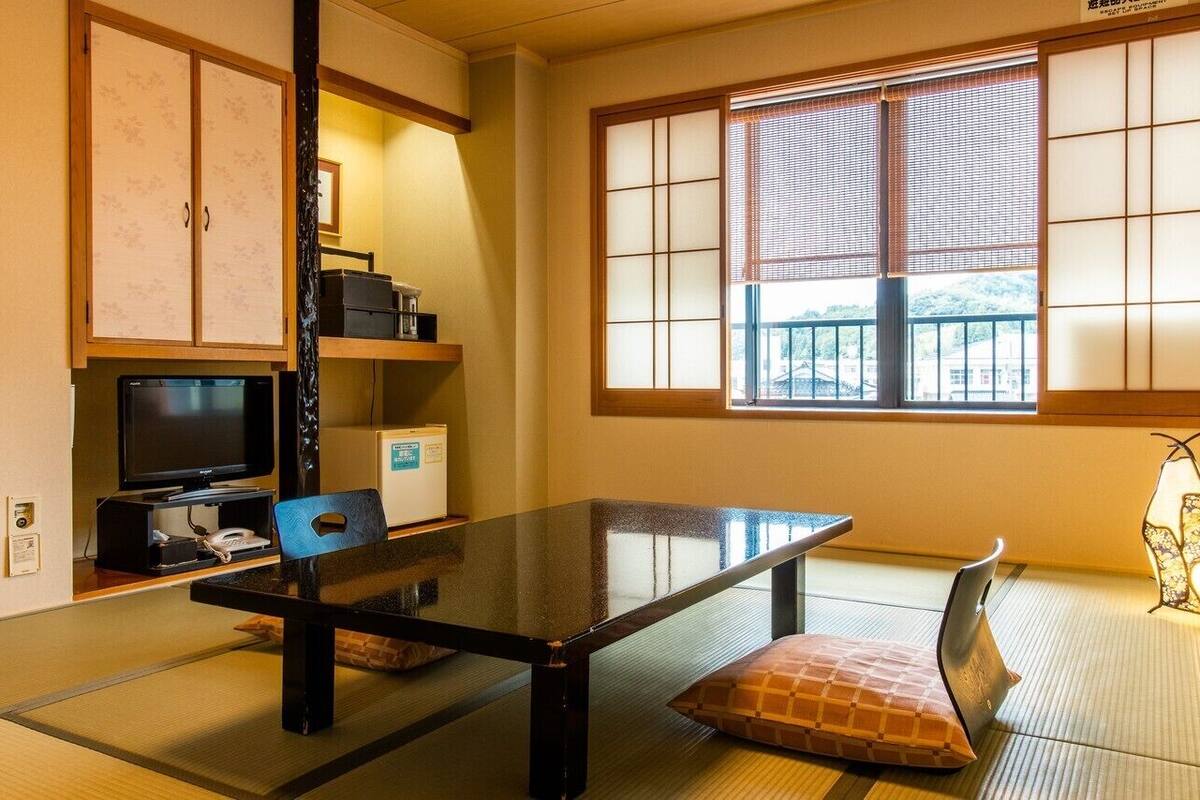 Une pièce de style japonais traditionnel, avec un sol recouvert de tatamis, une table basse, un téléviseur, un réfrigérateur et une fenêtre dotée de panneaux coulissants en papier.