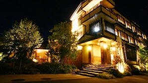 A multi-story hotel with a well-lit entrance, outdoor seating, and a staircase leading to the entrance. - Koujin no Yado Sanpou (Kami)