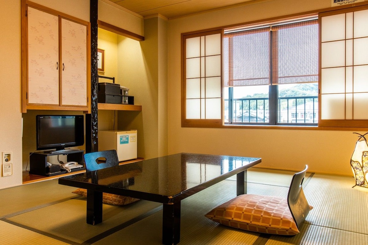A traditional Japanese room with tatami flooring, a low table, a television, a refrigerator, and a window with shoji screens.