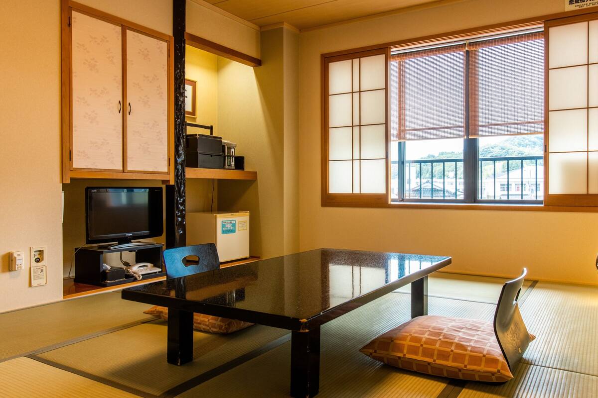 Une pièce de style japonais traditionnel, avec un sol recouvert de tatamis, une table basse, un téléviseur et une porte coulissante donnant sur un paysage.