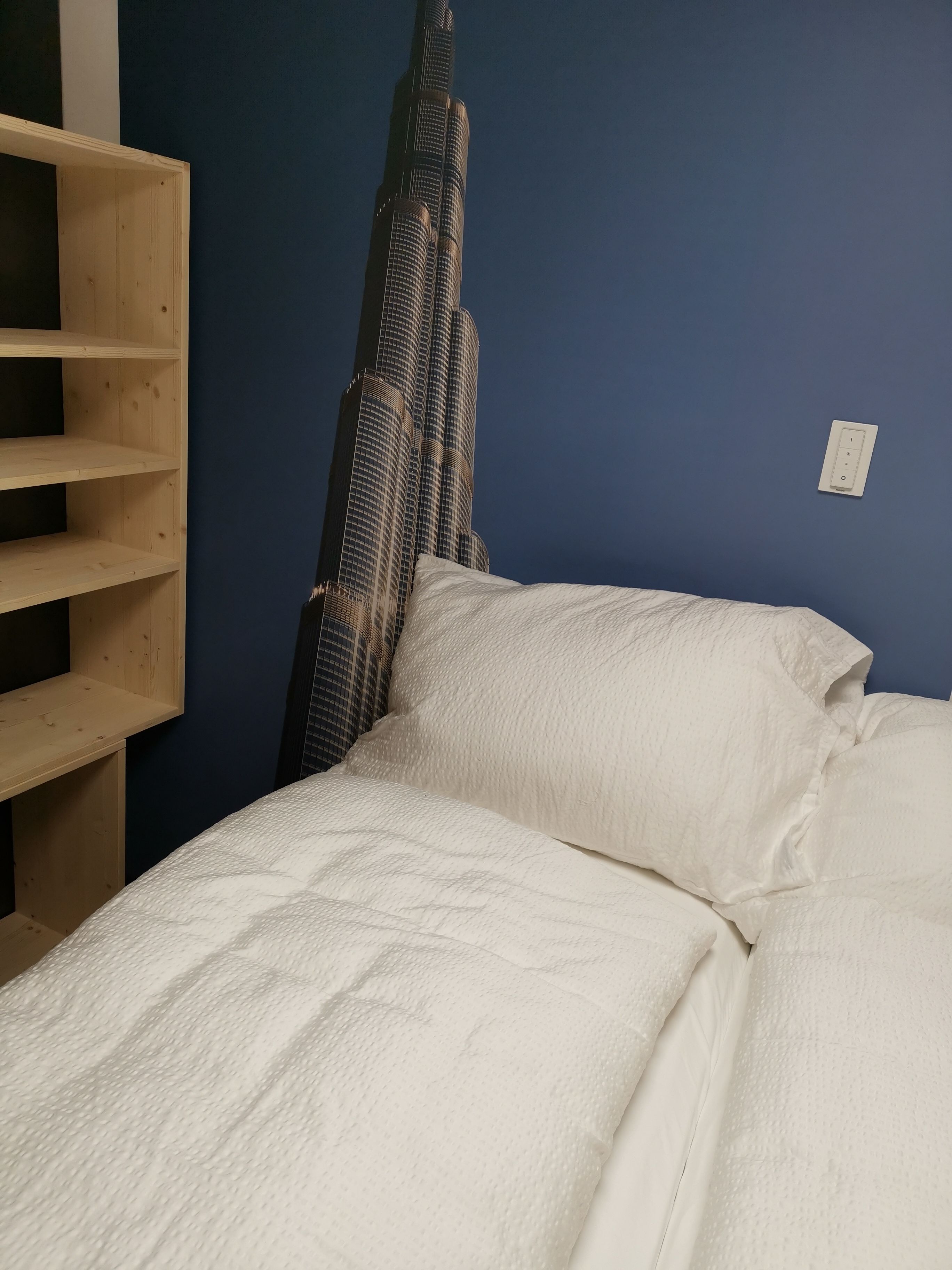 Una cama con ropa de cama blanca, una estantería de madera y un interruptor de luz en la pared.