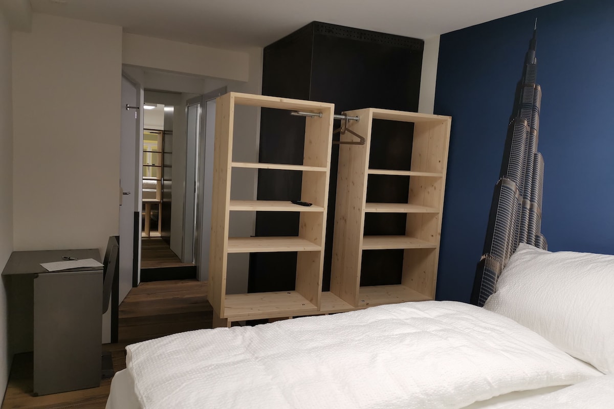 A bedroom with a bed, a wooden wardrobe, a desk, and a view of a tall building.
