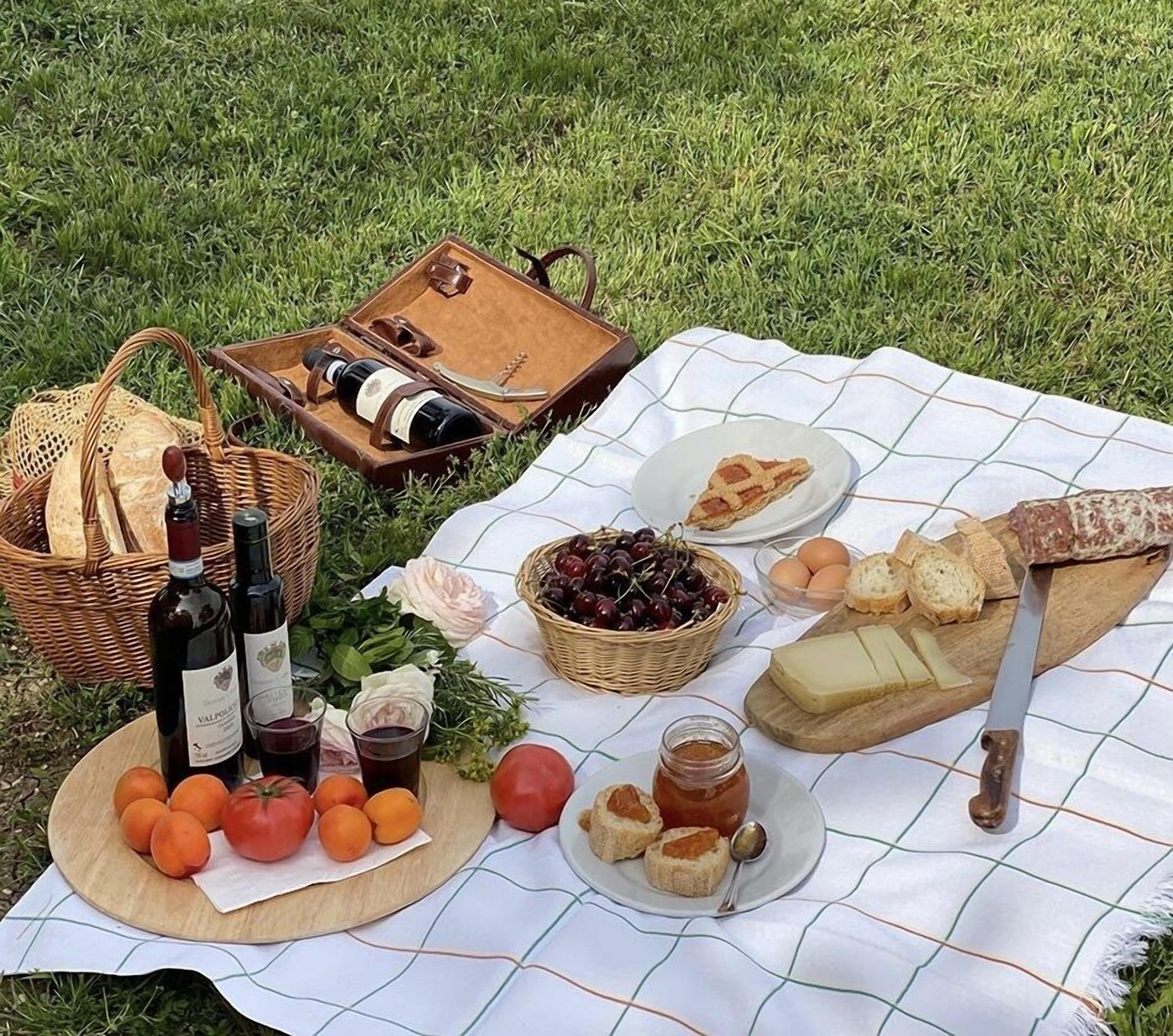 Un pique-nique installé sur l’herbe, avec une couverture, des bouteilles, des fruits, du fromage, du pain et un couteau.