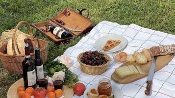 Un pique-nique installé sur l’herbe, avec une couverture, des bouteilles, des fruits, du fromage, du pain et un couteau.