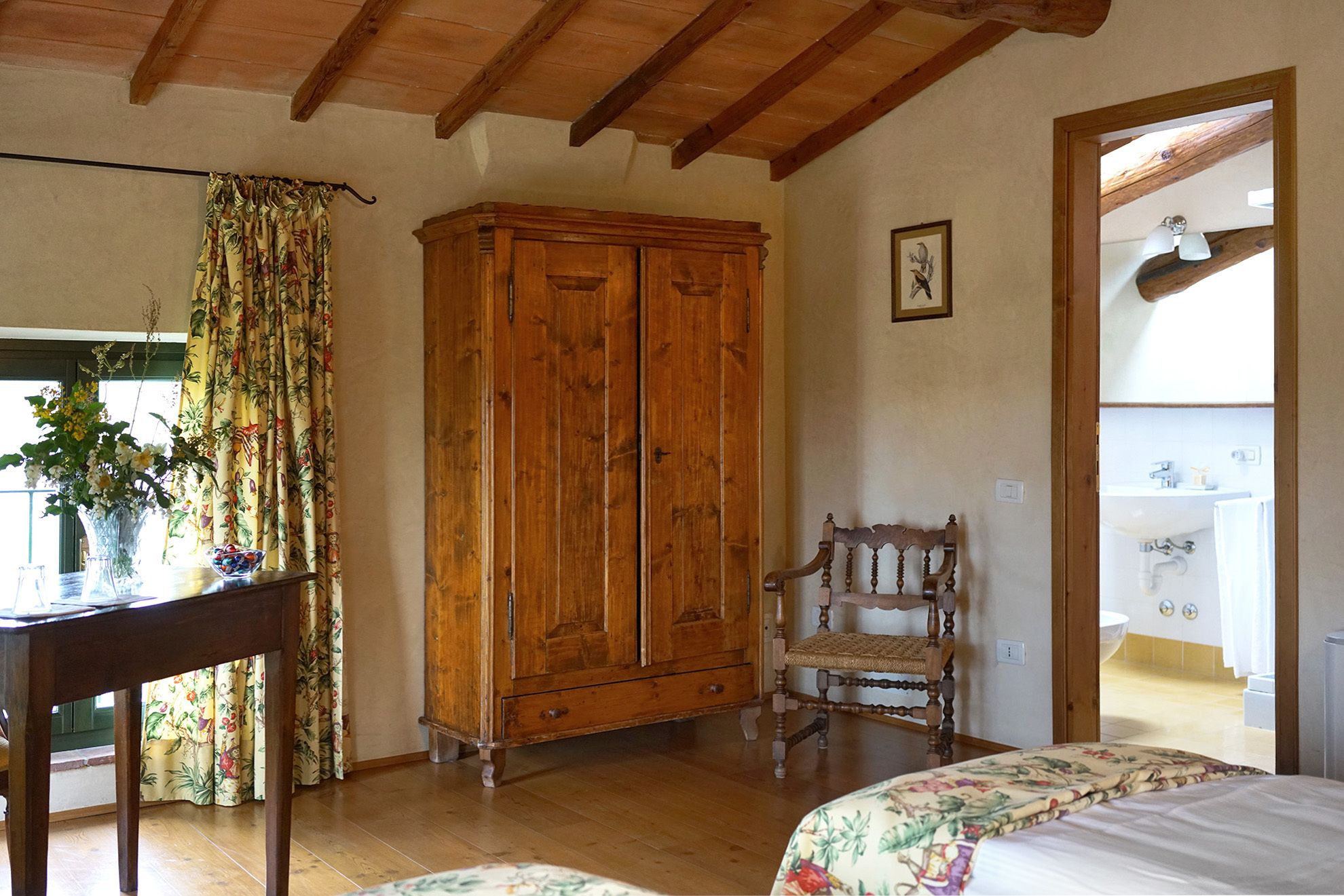 A room with wooden flooring, a wooden cabinet, a chair, a bed with floral bedding, and a window with curtains.