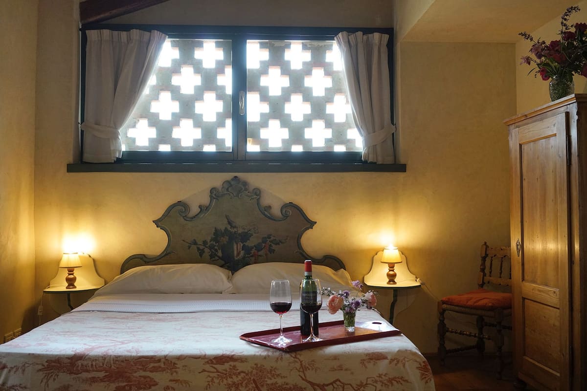 A hotel room with a bed, two bedside lamps, a window with a lattice design, a wooden headboard, a wooden cabinet, and a tray with a wine bottle and glasses.