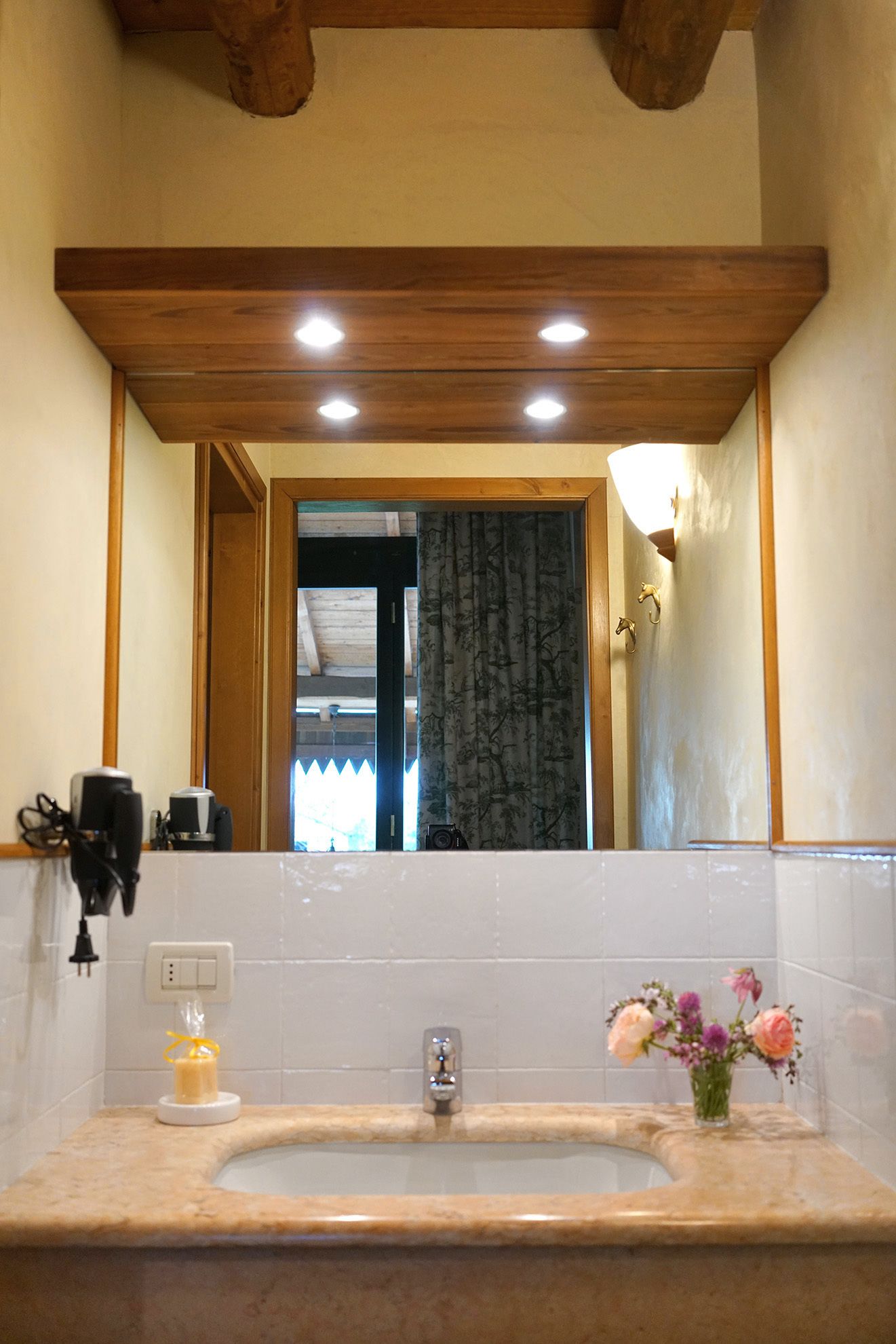 A bathroom with a wooden ceiling, a large mirror, a sink with a vase of flowers, and a view into the room.