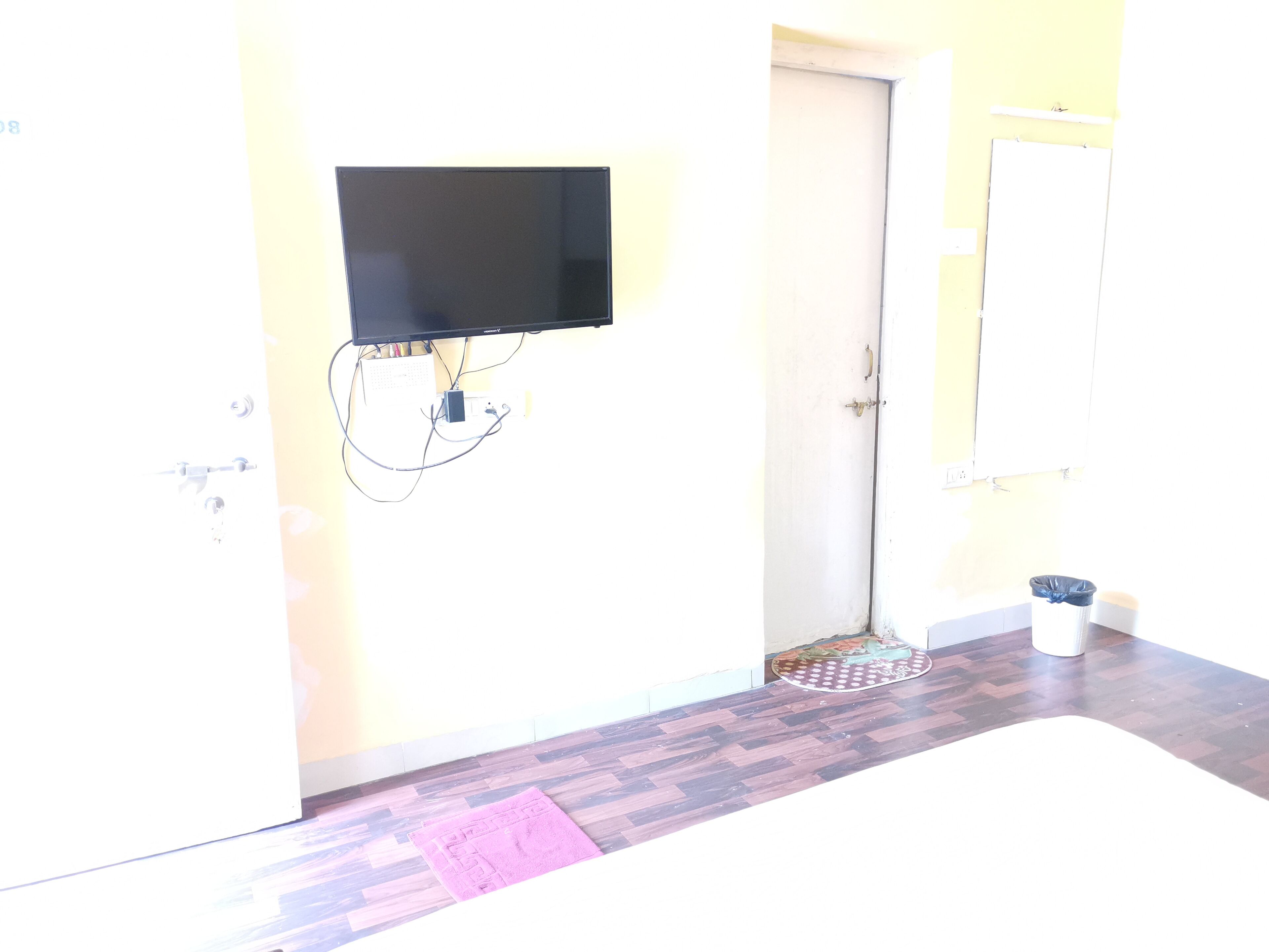 Une pièce dotée d’un téléviseur à écran plat fixé au mur, d’une porte, d’une petite poubelle et d’un tapis à motifs sur le sol.
