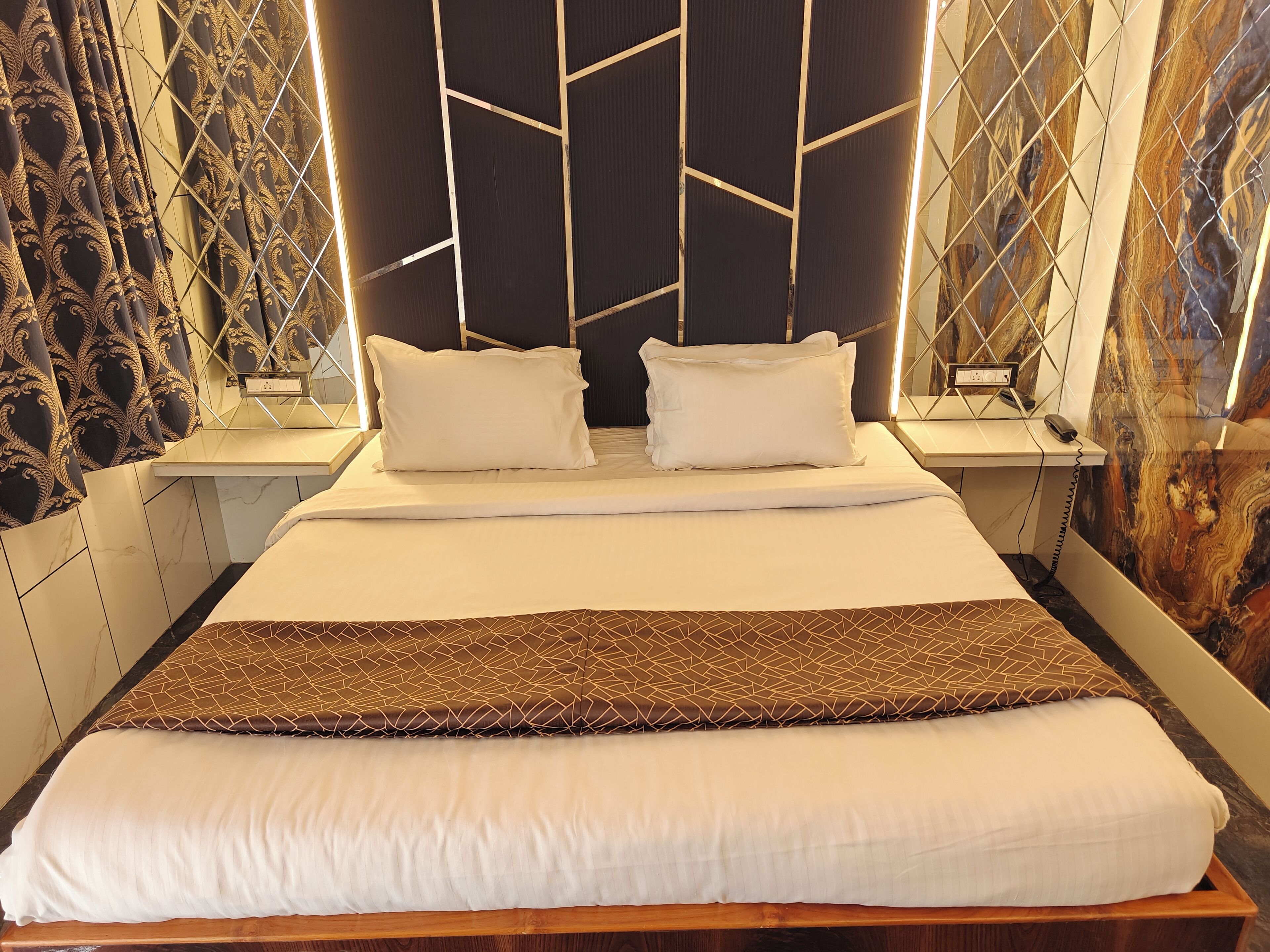 A neatly made bed with white linens and a brown decorative throw, a bedside table with a lamp, and a wall with a geometric pattern.