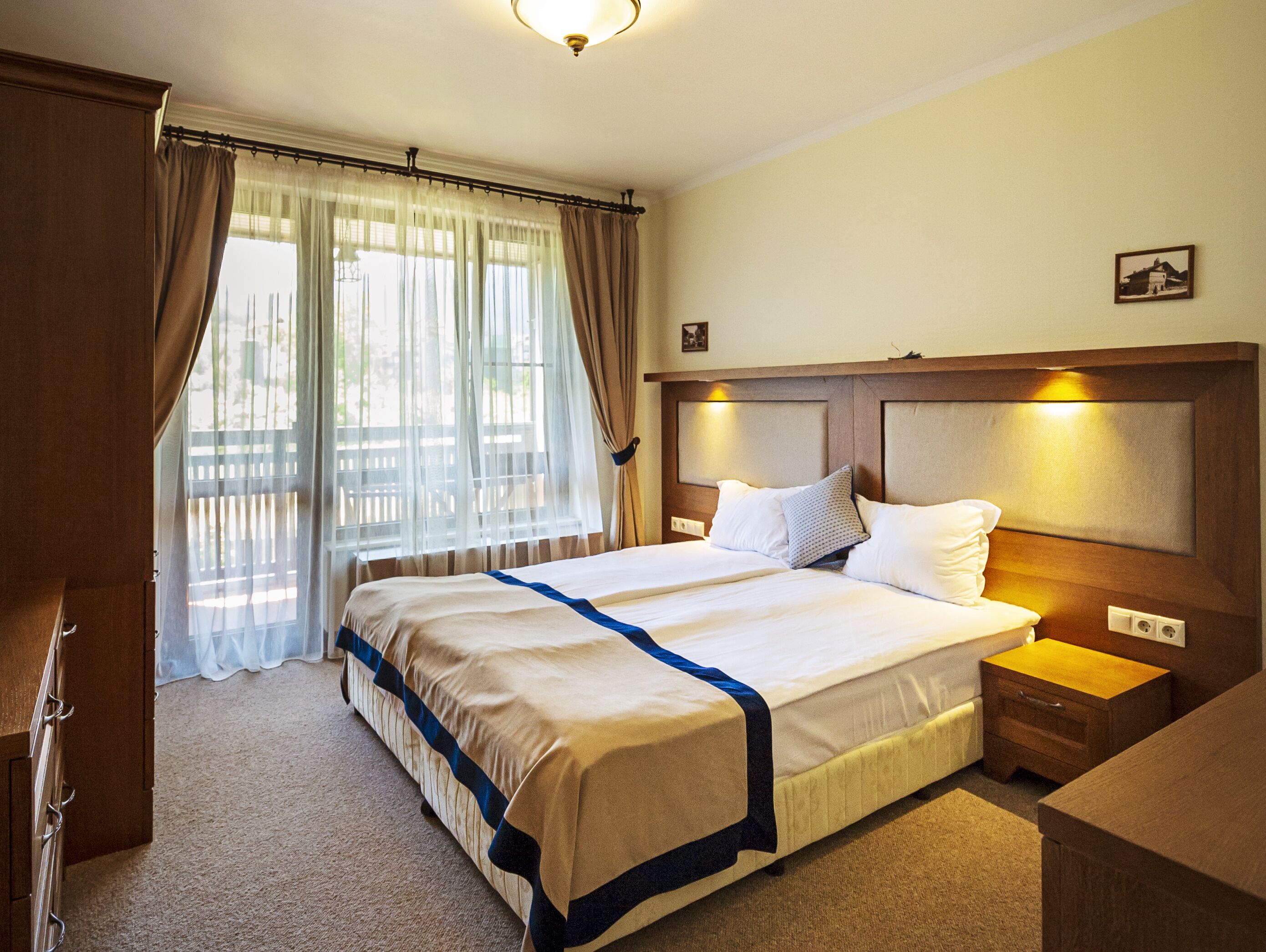 A hotel room with a large bed, a wooden headboard, a nightstand, a desk, and a window with curtains.