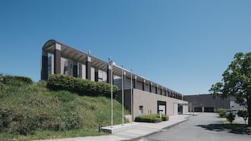 A modern building with a curved facade, a covered walkway, and a landscaped area with shrubs and a tree.
