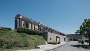 Un edificio moderno con fachada curva, un paseo techado y un área ajardinada con arbustos y un árbol.