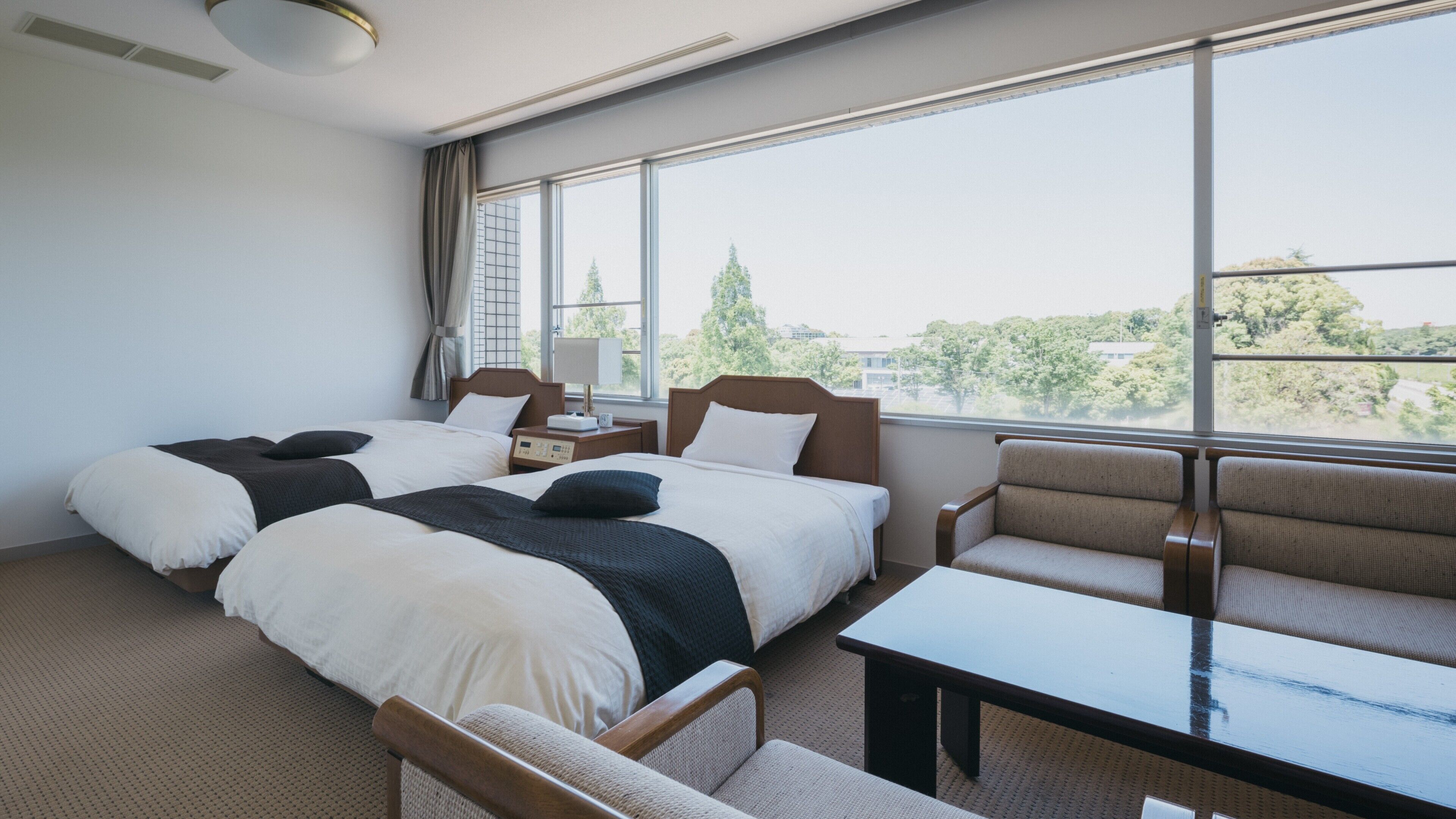 Ein Hotelzimmer mit zwei Betten, einem Schlafsofa, einem Tisch und großen Fenstern mit Blick auf Bäume und Gebäude.