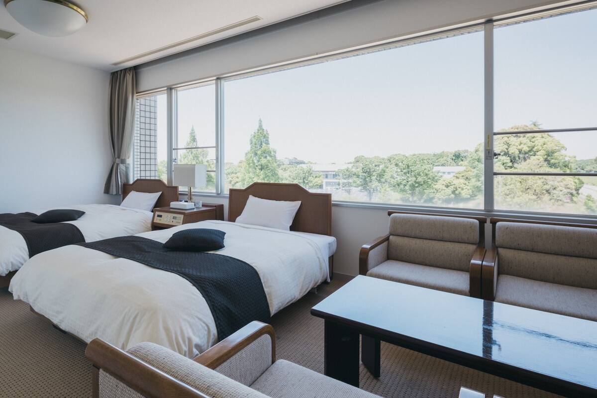 A hotel room with two beds, a sofa, a table, and large windows offering a view of trees and buildings.